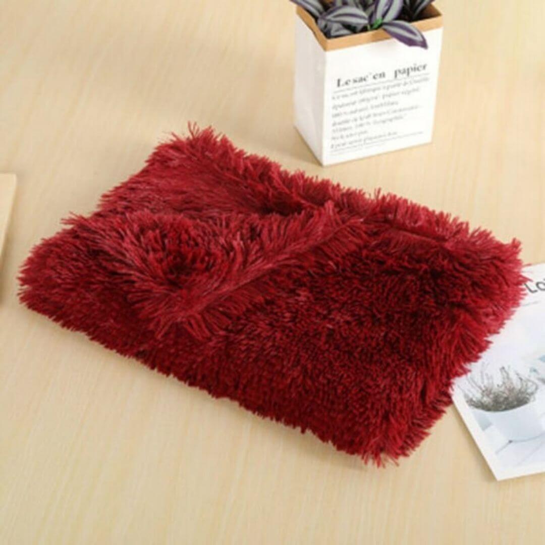 A soft wine red anxiety relieving blanket folded nicely on a wooden table with a little potted plant and some papers
